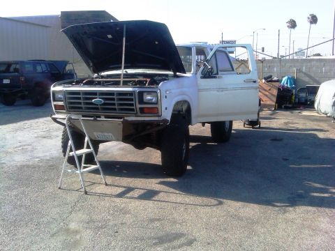 1986 Ford Bronco - White Ford truck with hood open and driver door ajar, parked outside with another vehicle in the background.