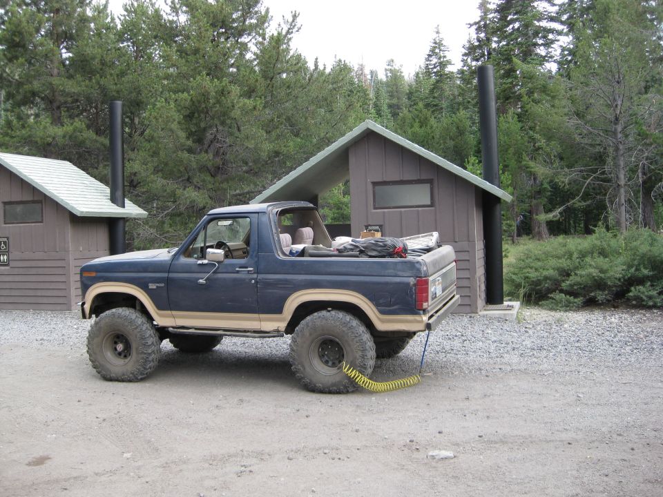 Blue Ford Bronco with large off-road tires parked next to a wooden outhouse