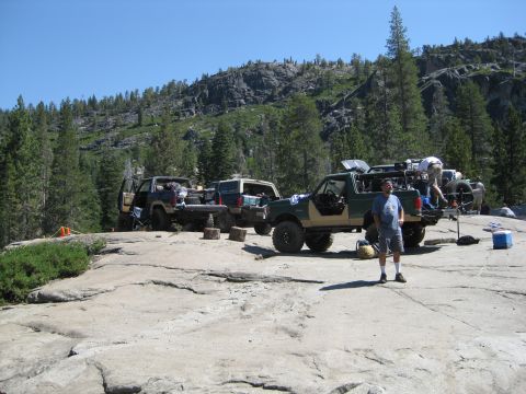 1986 Ford Bronco - A group of off-road vehicles are parked on a rocky outcrop, with people loading gear into a green and tan Ford Bronco.