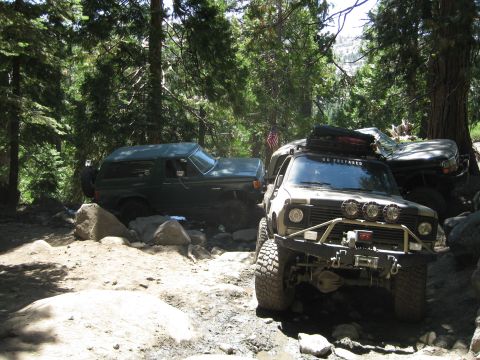 1986 Ford Bronco - Two off-road vehicles tackle a challenging rocky path surrounded by trees.