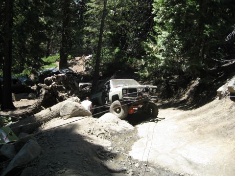 1986 Ford Bronco - A modified truck navigates a challenging off-road path through a wooded area, with another vehicle partially obscured in the background.