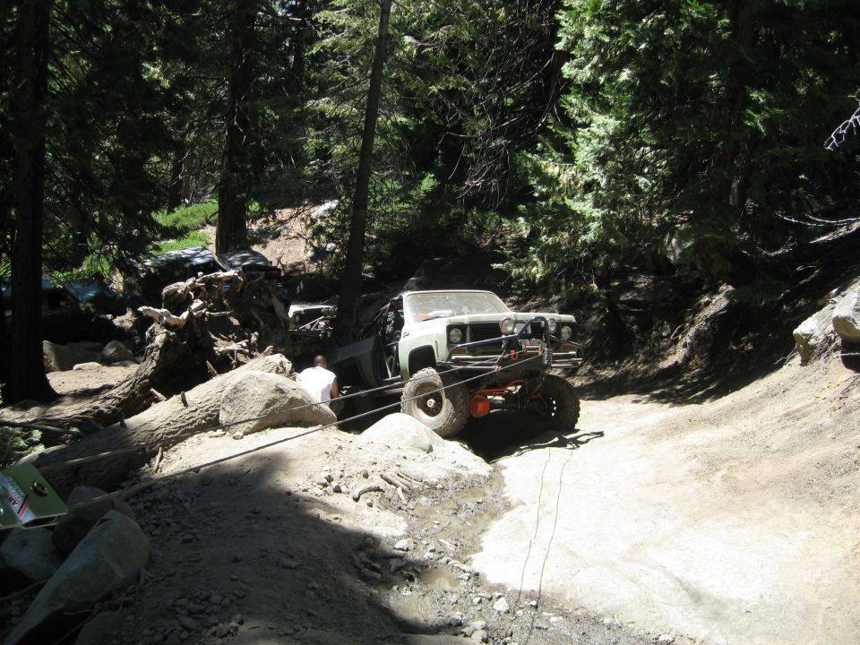 Off-road truck with lifted suspension and winch on a dirt trail, with another vehicle visible in the background among trees and rocks