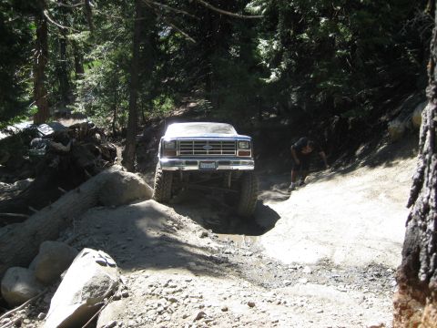 1986 Ford Bronco - A lifted 1980s Ford Bronco navigates a challenging off-road trail with a person observing nearby.