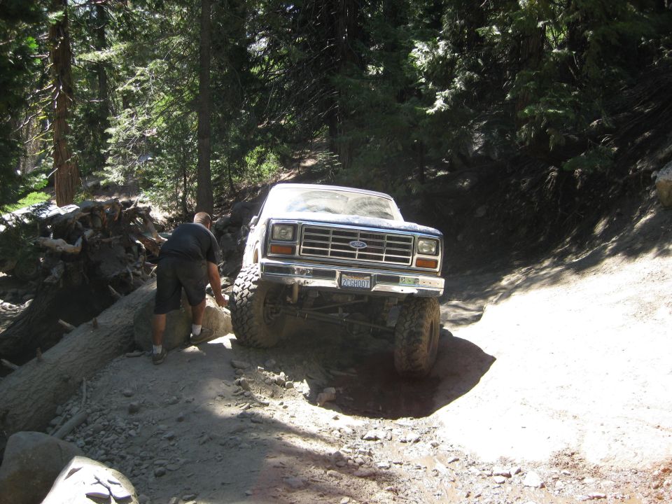 Ford Bronco with lifted suspension navigating a rocky, wooded trail. A person is outside the vehicle near the front tire