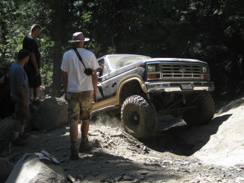 1986 Ford Bronco - This lifted 1986 Ford Bronco navigates a challenging off-road path with onlookers observing.