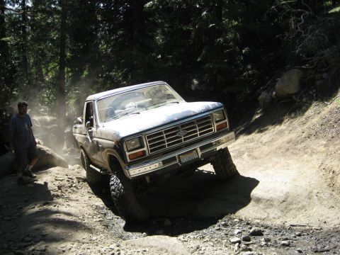 1986 Ford Bronco - This Ford pickup truck is navigating a challenging off-road trail, kicking up dust as it goes.