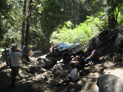1986 Ford Bronco - A group of people watch as a black pickup truck tackles a challenging rocky trail in a wooded area.