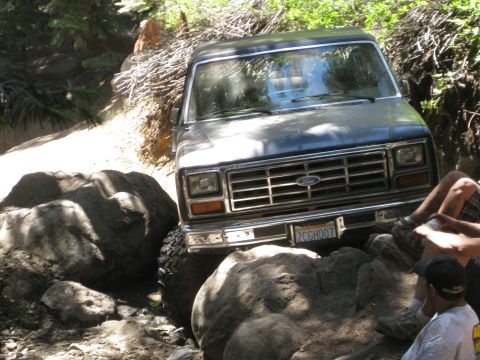 1986 Ford Bronco - A dark-colored Ford Bronco is shown navigating a rocky off-road trail with trees and brush surrounding it.
