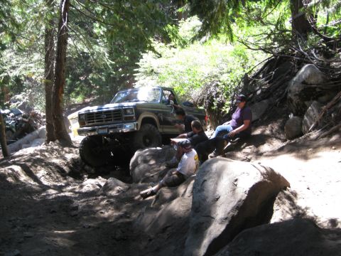 1986 Ford Bronco - A lifted 1980s Ford Bronco tackles a challenging off-road trail with passengers watching from the side.
