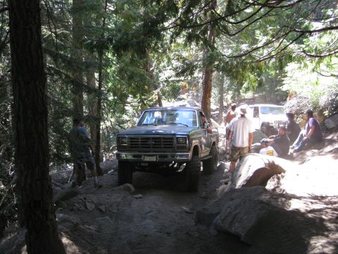 1986 Ford Bronco - A group of people are gathered around a lifted Ford pickup truck on a wooded, off-road trail.