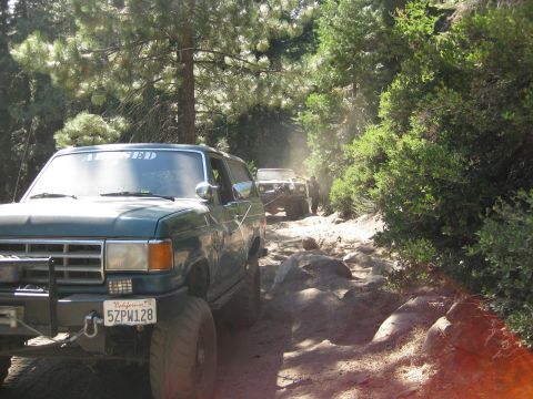 1986 Ford Bronco - Two off-road vehicles navigate a dusty, wooded trail with large rocks.