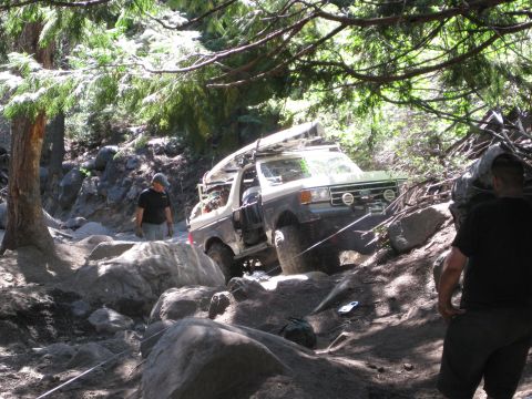1986 Ford Bronco - Ford Bronco tackling a challenging rocky trail in a wooded area, with two people observing.