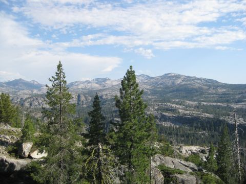 1986 Ford Bronco - A vast expanse of rocky mountains and evergreen forests stretches under a blue sky dotted with white clouds.