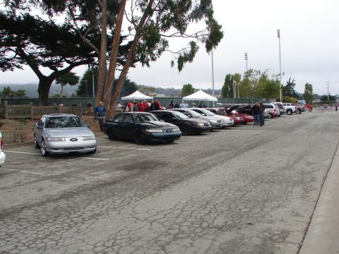 1992 Ford Taurus - A silver 1992 Ford Taurus is parked in a lot with other vehicles and tents in the background.