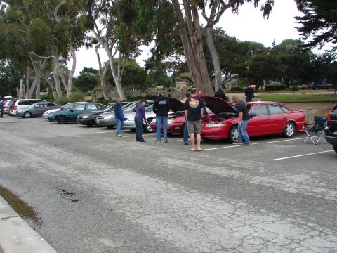 1992 Ford Taurus - Several cars are parked in a lot with people gathered around one with its hood open.