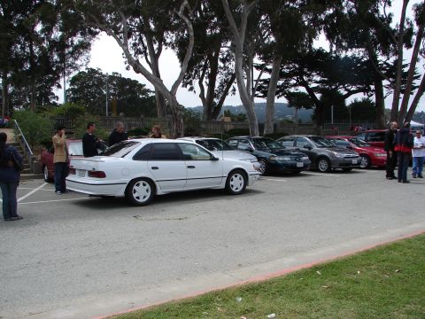 1992 Ford Taurus - A white 1992 Ford Taurus is parked in a lot with several other vehicles and people milling about.