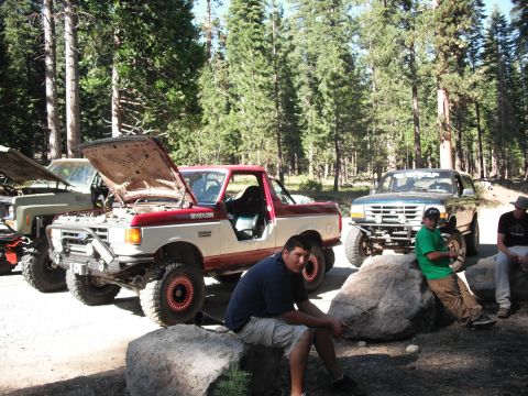 1991 Ford Bronco - A group of off-road vehicles are parked in a forest clearing, with several people relaxing on large rocks.