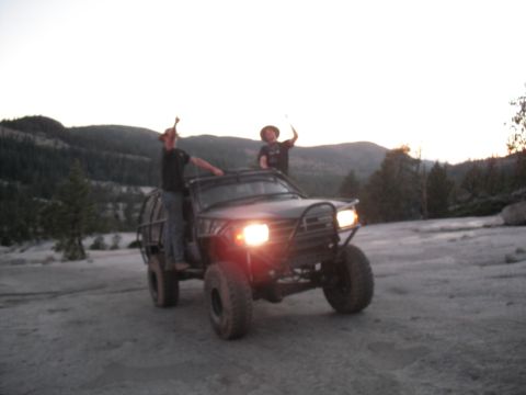 1991 Ford Bronco - Two people celebrate atop a heavily modified off-road truck in a rocky, wooded area at dusk.