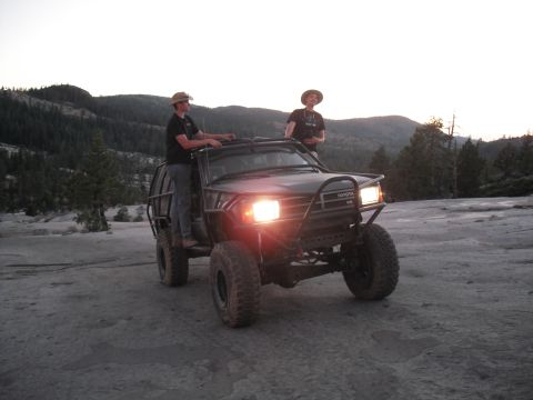 1991 Ford Bronco - Two individuals stand atop a modified Toyota off-road vehicle in a mountainous, rocky landscape at dusk.
