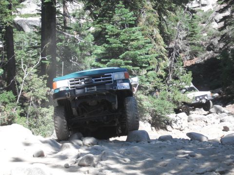 1991 Ford Bronco - Two off-road vehicles tackle a challenging rocky trail in a wooded area.