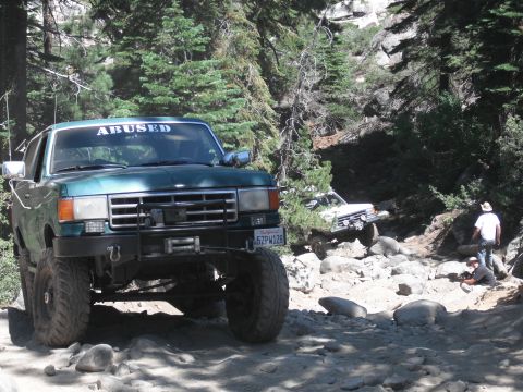 1991 Ford Bronco - A lifted green Ford Bronco navigates a challenging, rocky off-road path with another vehicle visible in the background.