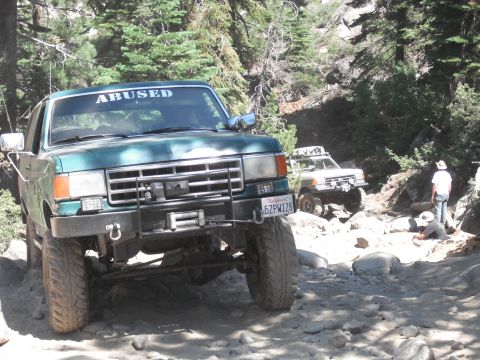 1991 Ford Bronco - A green 1991 Ford Bronco navigates a challenging off-road trail, with another vehicle visible behind it.