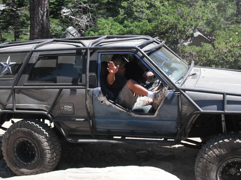 Gray off-road vehicle with external roll cage, driver's door removed, driver waving from interior