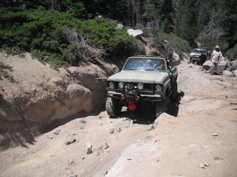 1991 Ford Bronco - Two off-road vehicles navigate a challenging, rocky path through a wooded area on a sunny day.