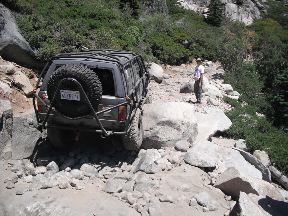 Gray off-road SUV with external roll cage and spare tire on rear, navigating a rocky trail