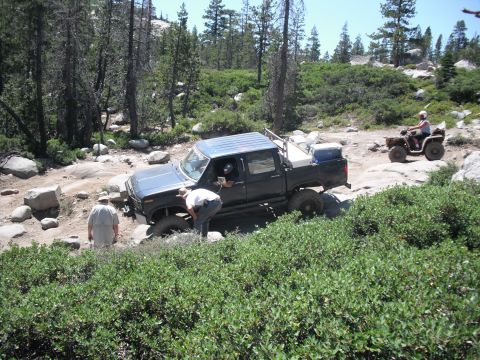1991 Ford Bronco - A dark blue pickup truck navigates a challenging rocky trail with one front tire lifted.
