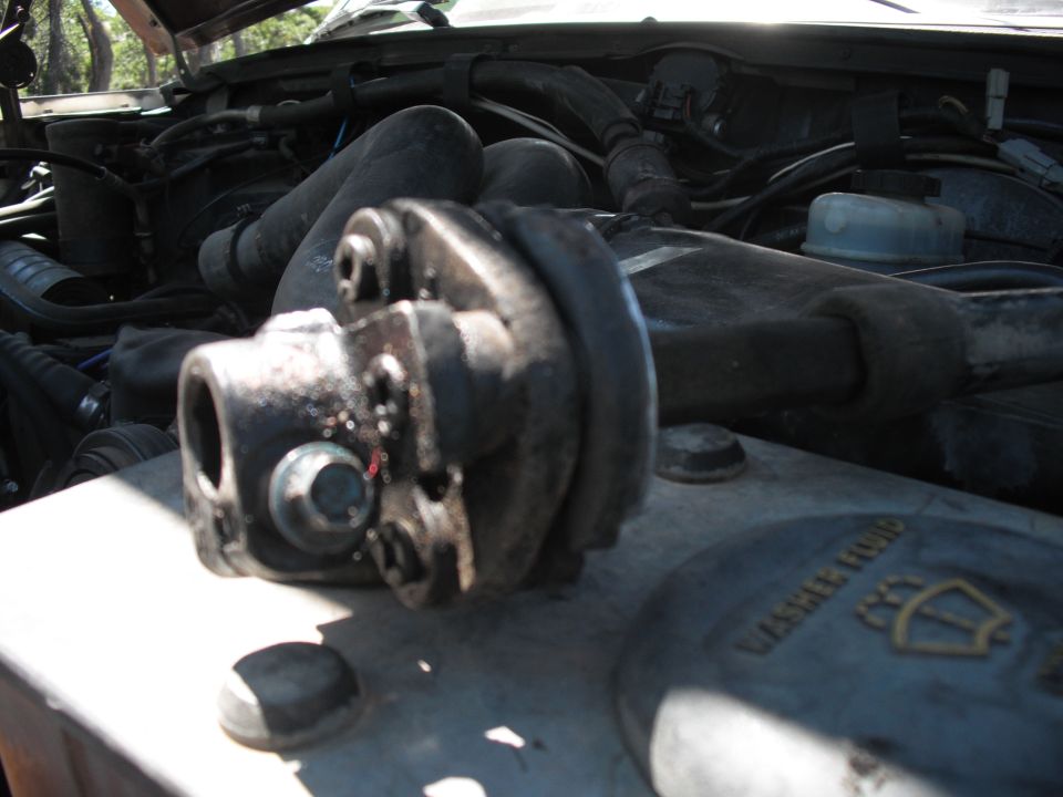Close-up of a greasy steering universal joint in a vehicle's engine bay, with hoses and engine parts visible