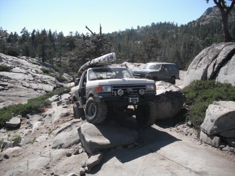 1991 Ford Bronco - Two off-road vehicles traverse a challenging rocky path in a mountainous, wooded area.