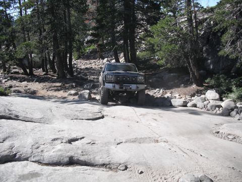 1991 Ford Bronco - This truck is navigating a rough, rocky terrain with trees and large boulders visible in the background.