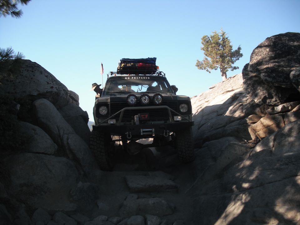 Off-road vehicle navigating a narrow, rocky path with a roof rack loaded with gear. Text on the windshield reads 'GO PREPARED'