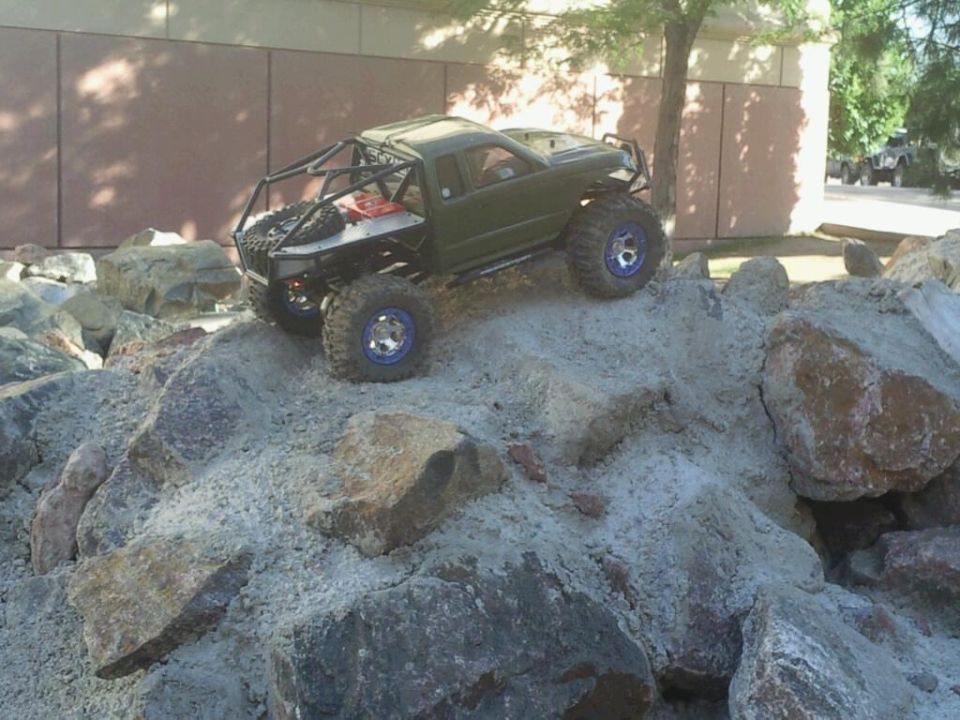 Olive-drab RC rock crawler pickup with tube cage and blue beadlock wheels on boulders