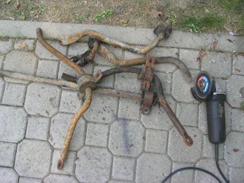 1986 Ford Bronco - Several heavily rusted sway bars lie on paver stones next to an angle grinder, suggesting removal or modification.