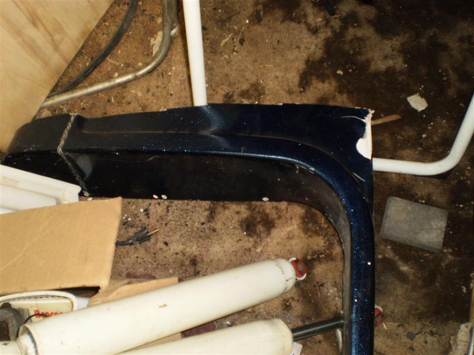 Dark blue fender flare with visible damage and white paint chips. Some white pipes and cardboard boxes are in the background