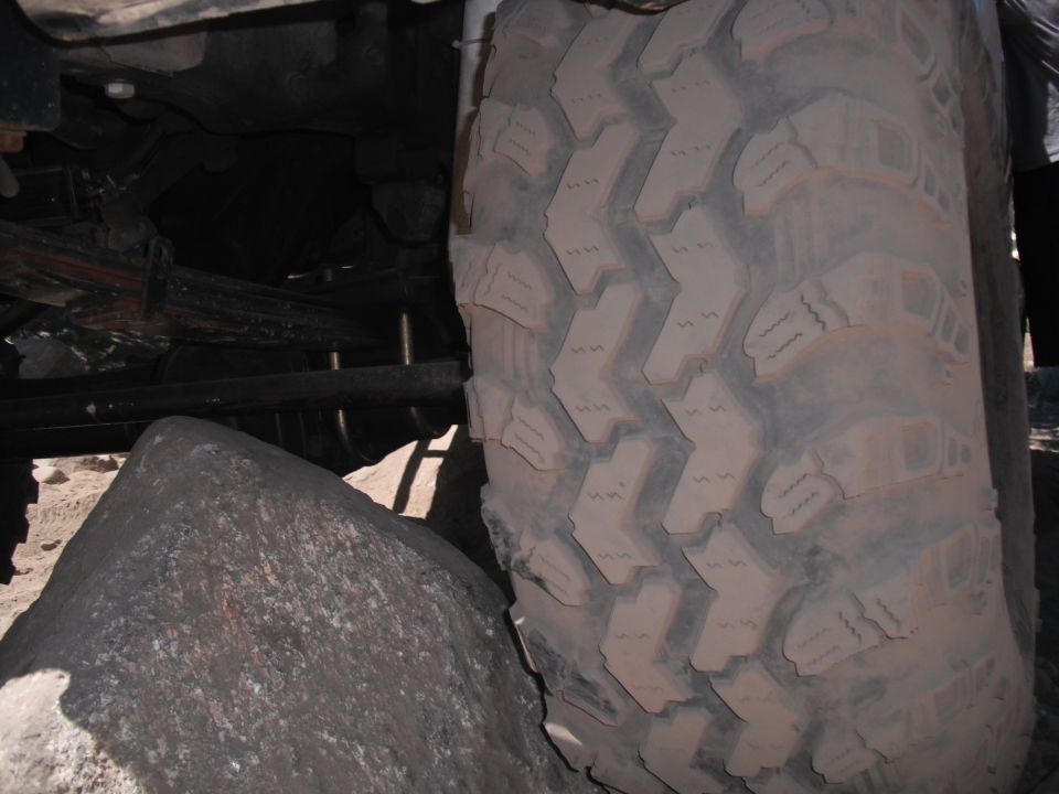 Close-up of a large off-road tire with aggressive tread, showing suspension parts and rocks