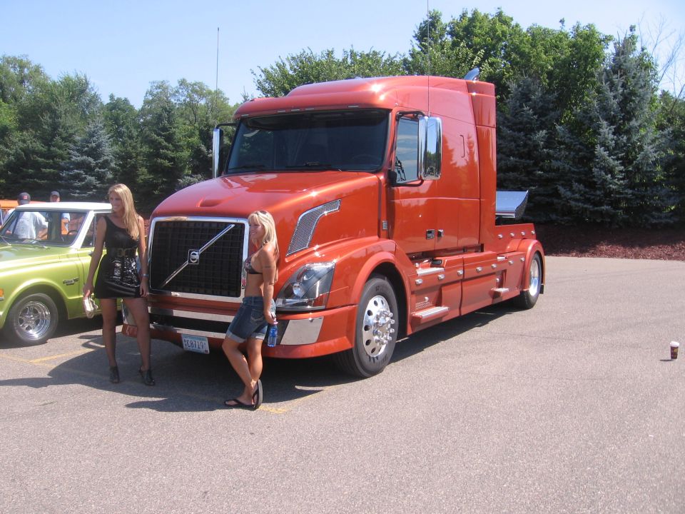 Orange semi-truck with Volvo grille, two women standing nearby, parked on asphalt