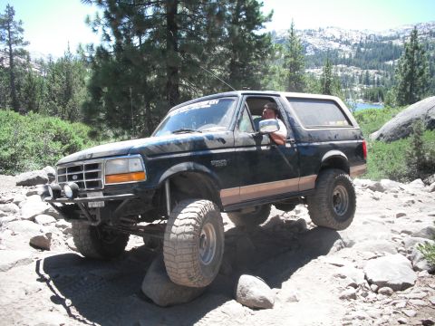 1991 Ford Bronco - A dark-colored Ford Bronco navigates a rocky off-road path with a lake and mountains in the background.