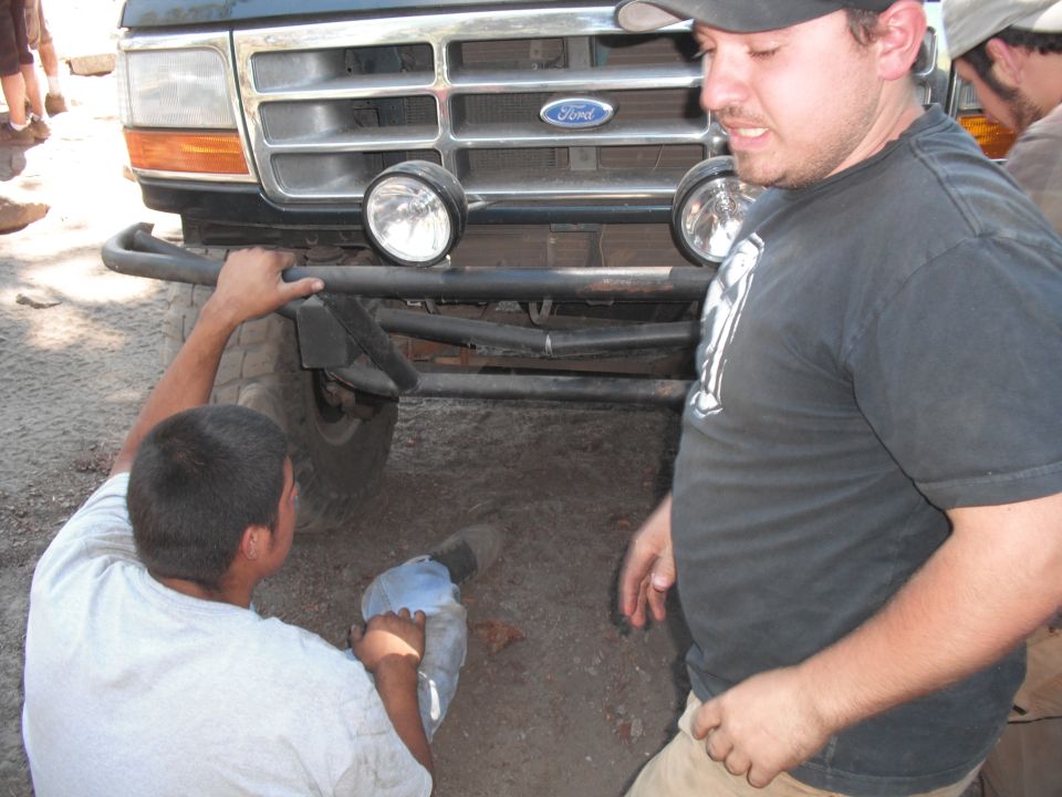 People working on the front bumper and grille of a Ford truck. The Ford logo is visible on the grille
