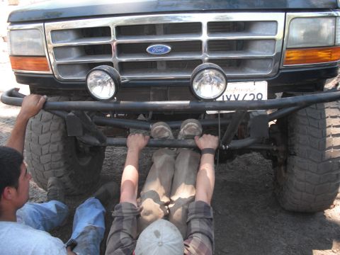 1991 Ford Bronco - Two people work on the front bumper and grille of a 1992 Ford Bronco, which has an aftermarket off-road bumper and auxiliary lights.