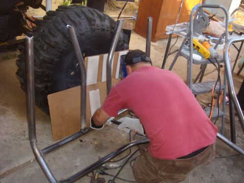 1992 Ford Bronco - A man welds a roll cage around a massive tire, with tools and a ladder visible in the background.