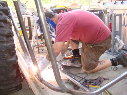 1992 Ford Bronco - A person welds metal tubing for a roll cage, with a large tire visible to the left.