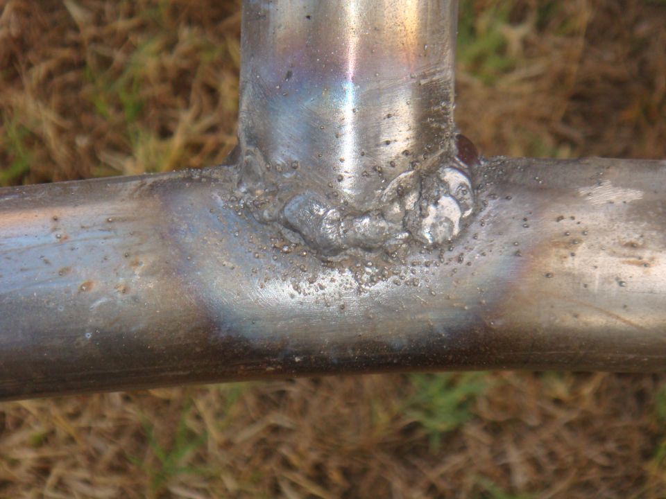 Welded metal tubing with rough welds and discoloration against a blurred grassy background