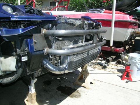 1995 Ford Probe - The front end of a blue 1995 Ford Probe is elevated on jack stands, revealing custom intercooler piping and a roll cage structure.