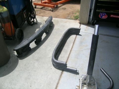1995 Ford Probe - Car parts, including a bumper and roll cage tubing, are laid out on a concrete surface next to a welding machine.