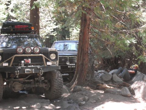 1991 Ford Bronco - Two off-road vehicles are parked in a forest clearing, with one person resting nearby.