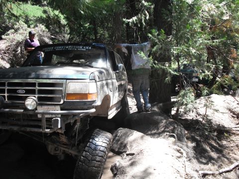 1991 Ford Bronco - A dark blue Ford Bronco navigates a challenging off-road trail with a "UNDER CONSTRUCTION 4X4" sticker on its windshield.
