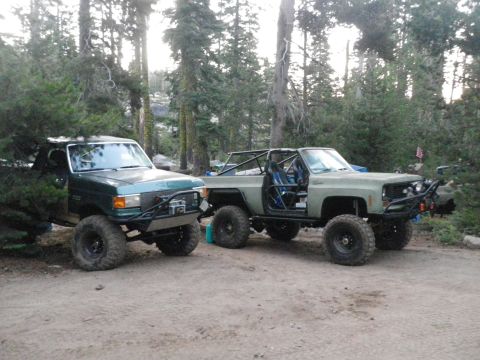 1991 Ford Bronco - Two heavily modified SUVs, one appearing to be a 1991 Ford Bronco, are parked on a dirt road surrounded by trees.